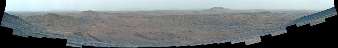 A rover traversing the red rocky surface of Mars with layered terrain and a dusty horizon behind it.
